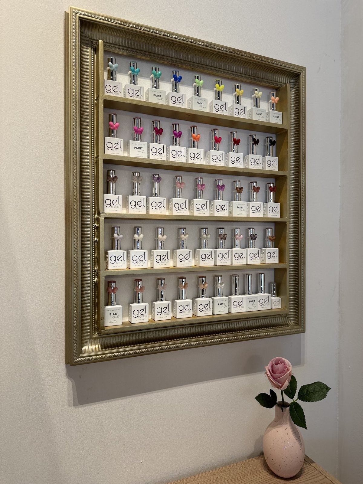 Gouden lijst aan de muur met rijen flesjes gellak, met een roos in een vaasje eronder